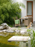 Blick vom Schwimmteich mit Holzsteg auf Terrasse mit Korbmöbeln