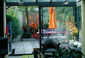 Blick aus der Wohnung mit blauen Caféstühlen, pinkfarbenem Holztisch, orangefarbenem Sonnenschirm, Terrassenbelag, orangefarbener Canna und verzinktem Tablett mit Sukkulentenbepflanzung