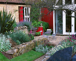 Blick auf die Rückseite des Hauses mit roter Wand, Stachys, Phormium, Metalltisch und Stühlen.