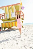 A brunette woman wearing a pink dress, statement necklace and sneakers in front of a lifeguard tower on the beach