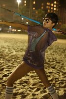 Dunkelhaarige Frau in türkisfarbenem Badeanzug, Glitzer-Shorts und Netzoberteil beim Speerwerfen am Strand