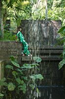 Brünette Frau in Hosenanzug mit tropischem Print, Sandalen und Sonnenbrille hinter einem Wasserfall