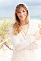 A blonde woman wearing a cream jumper and a white hooded cardigan on the beach