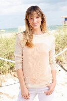A blonde woman wearing a cream jumper and white trousers on the beach