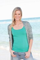 A blonde woman wearing a green top, grey cardigan and jeans on the beach