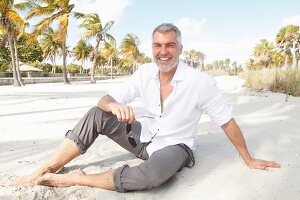 Mann mit grauen Haaren in weißem Hemd und grauer Hose sitzt am Strand
