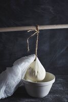 Ricotta in a cheesecloth hanging from a wooden stick and draining into a bowl