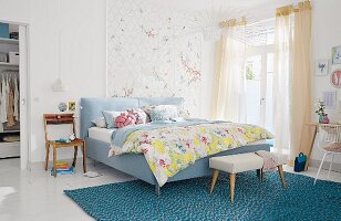 A padded double bed and bench on a turquoise rug in a light, bright bedroom