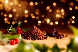 Ilex auf Holztisch mit einem Haufen Kakaopulver, Kakaobohnen und Schokoröllchen (weihnachtlich)