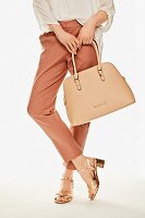 A woman wearing rust-coloured trousers and metallic-look sandals, carrying a nude-coloured handbag