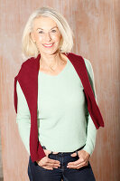 An older blonde woman wearing a light green knitted jumper with a dark red jumper over her shoulders
