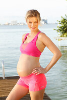 A pregnant woman on a jetty wearing a pink sports bra and shorts