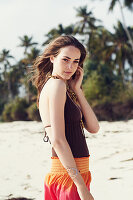 A young brunette woman on a beach wearing a brown bathing suit and harem trousers