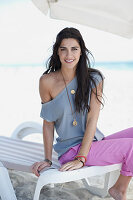 A young, long-haired woman on a lounger on a beach wearing a grey blouse and pink trousers