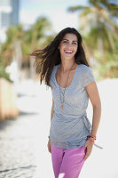 A young, long-haired woman on a lounger on a beach wearing a grey blouse and pink trousers