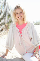 A young blonde woman sitting on a beach wearing a white blouse over a pink top