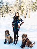 A young woman wearing a dress and a cardigan with two dogs in the snow