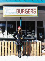 A young woman outside a burger restaurant wearing a dark jacket, a skirt, tights and boots