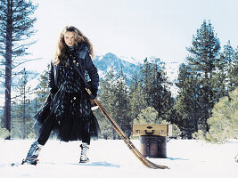 Junge Frau im Kleid und Mantel in blauen Tönen mit Eishockeyschlägern im Schnee