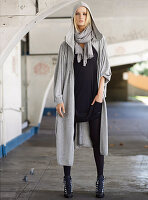 A young woman wearing a grey hooded coat, a black t-shirt dress and black tights