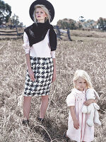 A blonde woman wearing a black hat, a light jumper and a skirt with a little girl in an autumnal field