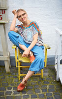 Blonde Frau mit Brille in Oversize-Top und Denim-Culotte auf Vintage Gartenstuhl sitzend