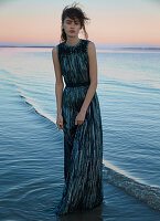 A dark-haired woman wearing a long evening dress by the sea at sunset