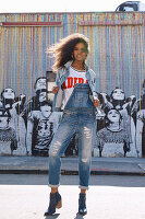 A dark-haired woman wearing denim dungarees and a denim jacket standing against a graffitied wall