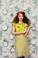 A dark-haired woman wearing a yellow blouse and a matching skirt standing against a papered wall hung with decorative plates