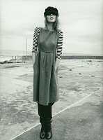 A young woman wearing a flat cap, a striped shirt and a dress by the sea