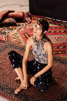 A young woman wearing a Paisley patterned summer blouse and black floral patterned trousers sitting on a rug