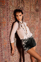 A young woman wearing a white blouse and black shorts standing against a hanging rug