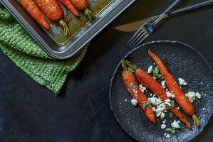 Roasted carrots with feta cheese on a black plate and in a roasting pan, on a black background