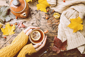 Cup of hot tea with lemon in woman's hands holding it over wooden autumn background