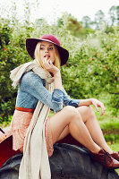 A blonde woman wearing a hat and a scarf, a denim blouse and a skirt sitting on a tractor