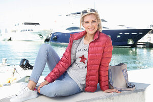 A blonde woman wearing jeans, a top and quilted jacket sitting at a harbour