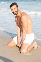 A young topless man kneeling in the sand wearing shorts