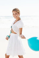 Blonde Frau mit blauem Luftballon in weißem Bolero und Rock mit Stickerei am Strand