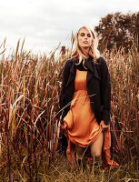 Blonde Frau in orangefarbenem Kleid und dunklem Mantel auf dem Getreidefeld