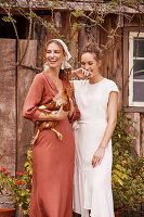 A woman wearing a headscarf and a brown dress holding a hen with her friend standing next to her wearing a white summer dress