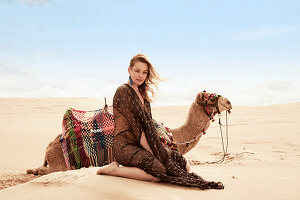 A young woman wearing a light, brown summer dress sitting next to a camel in the sand