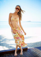 A young woman on a beach wearing a bikini top and a floral patterned skirt