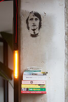 Stacked books on floating shelf, lamp and stencil portrait on wall