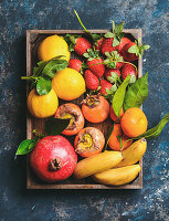 Healthy fresh fruit variety: Oranges, lemons with leaves, pomegranate, bananas, strawberries and persimmon in wooden box