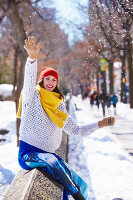 A young woman wearing a red hat, a yellow scarf, a white jumper and blue trousers in a snowy park