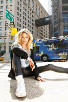 A young blonde woman wearing a black cardigan and faux leather trousers sitting on a pavement