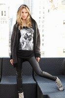 A young blonde woman wearing a t-shirt, a black hoodie, black trousers and leopard-print ankle boots