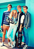 A group of young people wearing fashionable clothing posing against a wall with skateboards