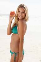 Reife blonde Frau mit Graperfuithälfte in türkisfarbenem Bikini am Strand