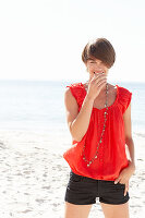 A mature brunette woman on a beach wearing a red shirt, a necklace and black shorts